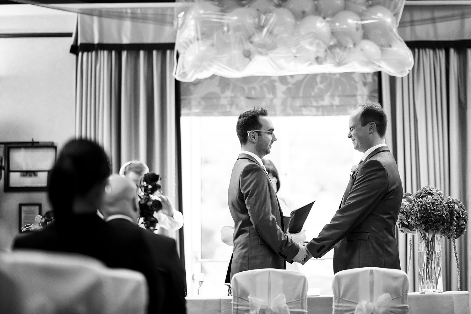 Two people hold hands during an intimate indoor wedding ceremony under decorative white clouds.