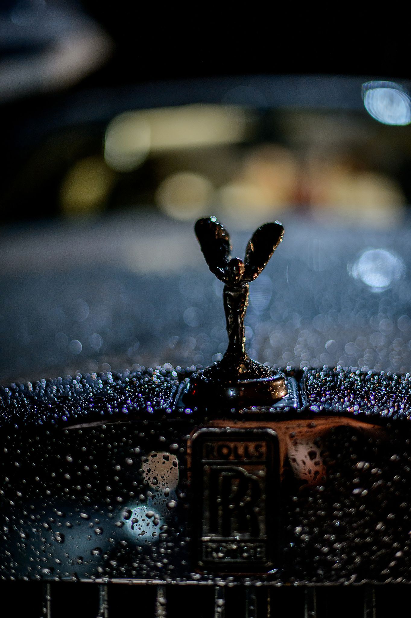 Close-up of a wet Rolls-Royce hood ornament at night with bokeh lights in background.