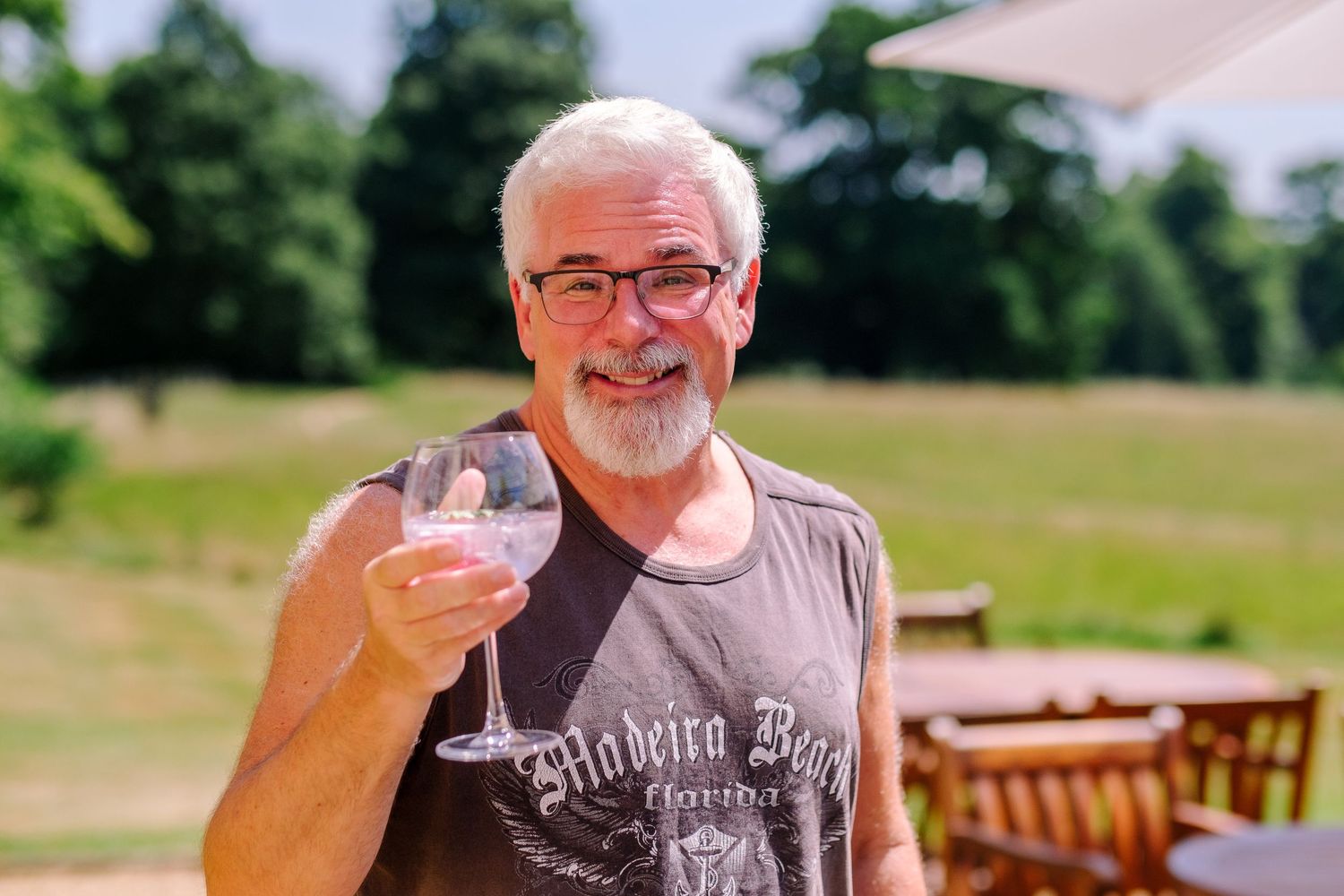 A person enjoying a drink outdoors on a sunny day at a wooden table in a garden setting.