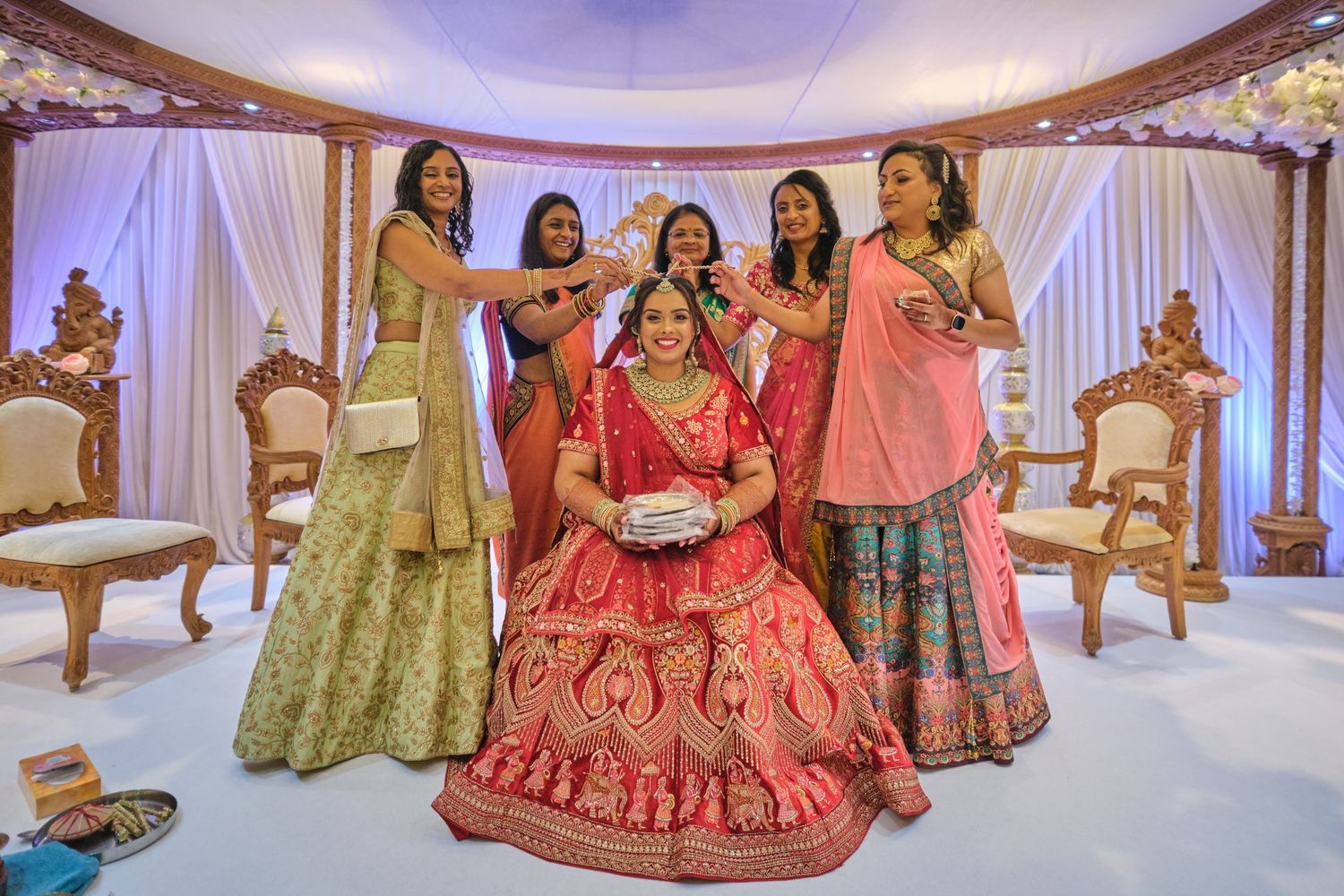 A group in traditional Indian wedding attire poses together on an elegant stage with white drapes and ornate chairs.