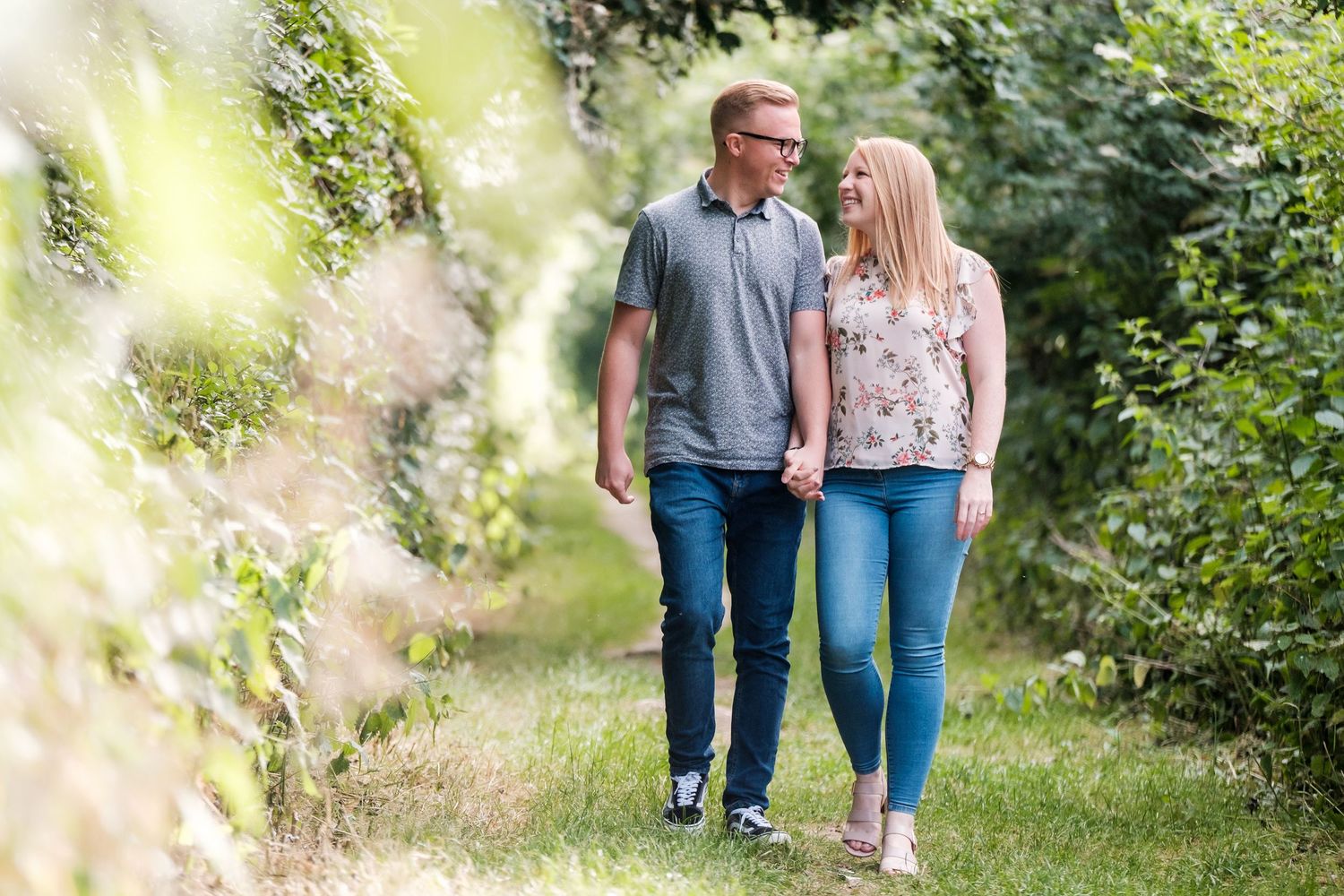 A couple walks together through a lush garden path surrounded by greenery on a sunny day.