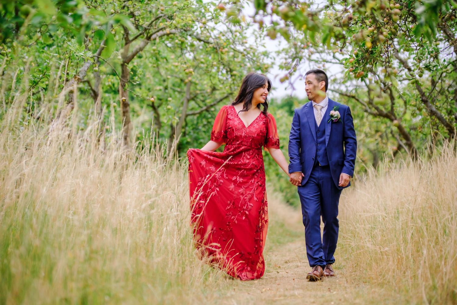 A couple in formal attire walks through tall grass and greenery in a romantic outdoor photo series.