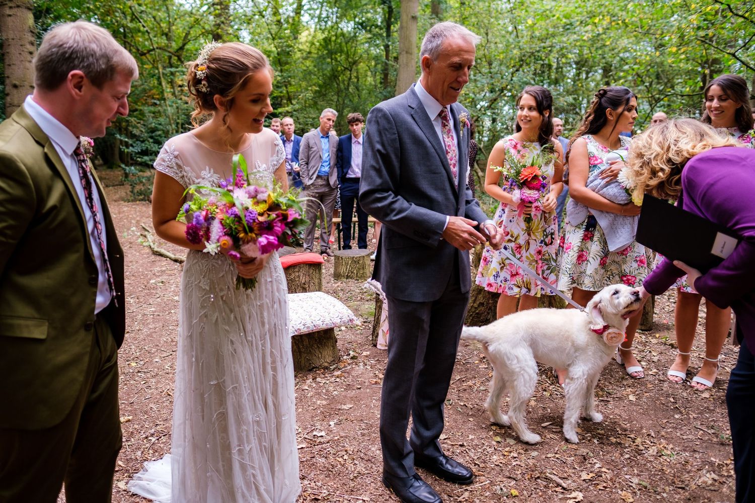 Outdoor wedding ceremony in a wooded setting with a white dog present.