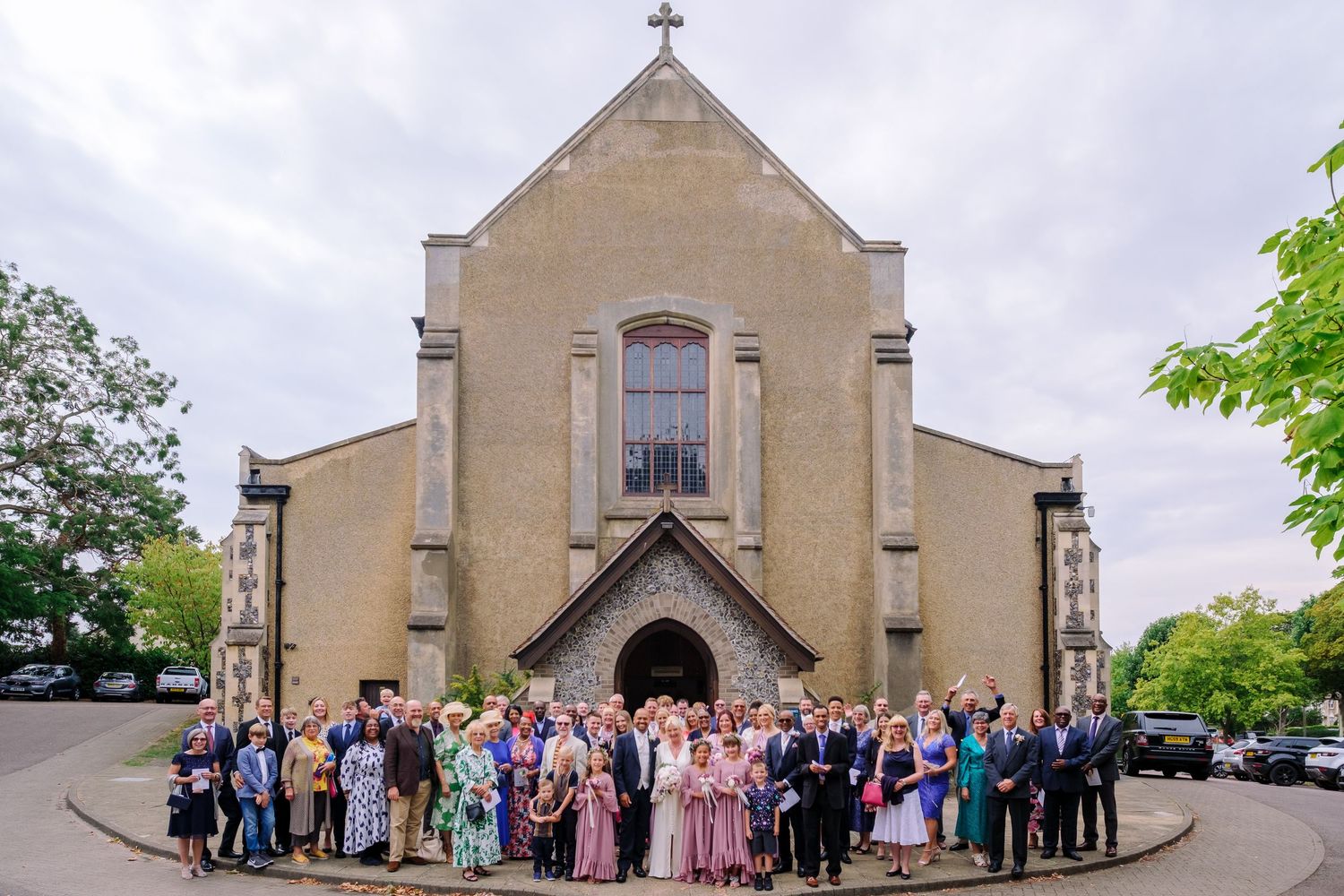 A panoramic view of a stone church exterior with wedding guests gathered outside on a sunny day.