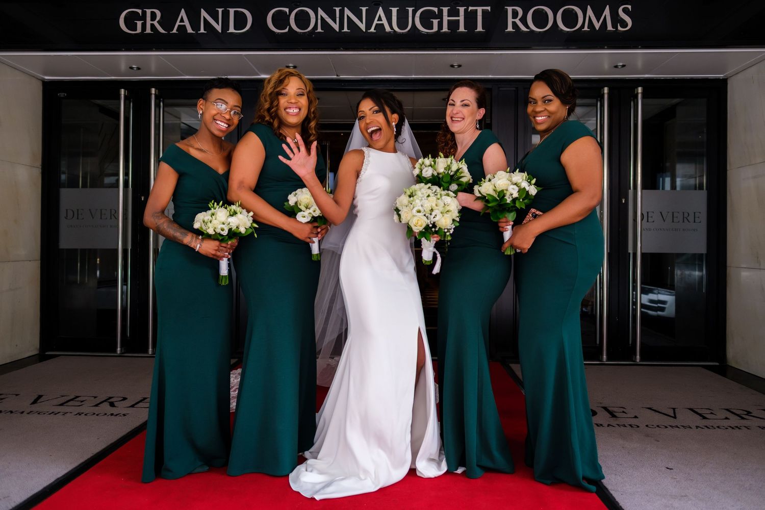 Wedding party in emerald green dresses and white gown pose together at Grand Connaught Rooms entrance.