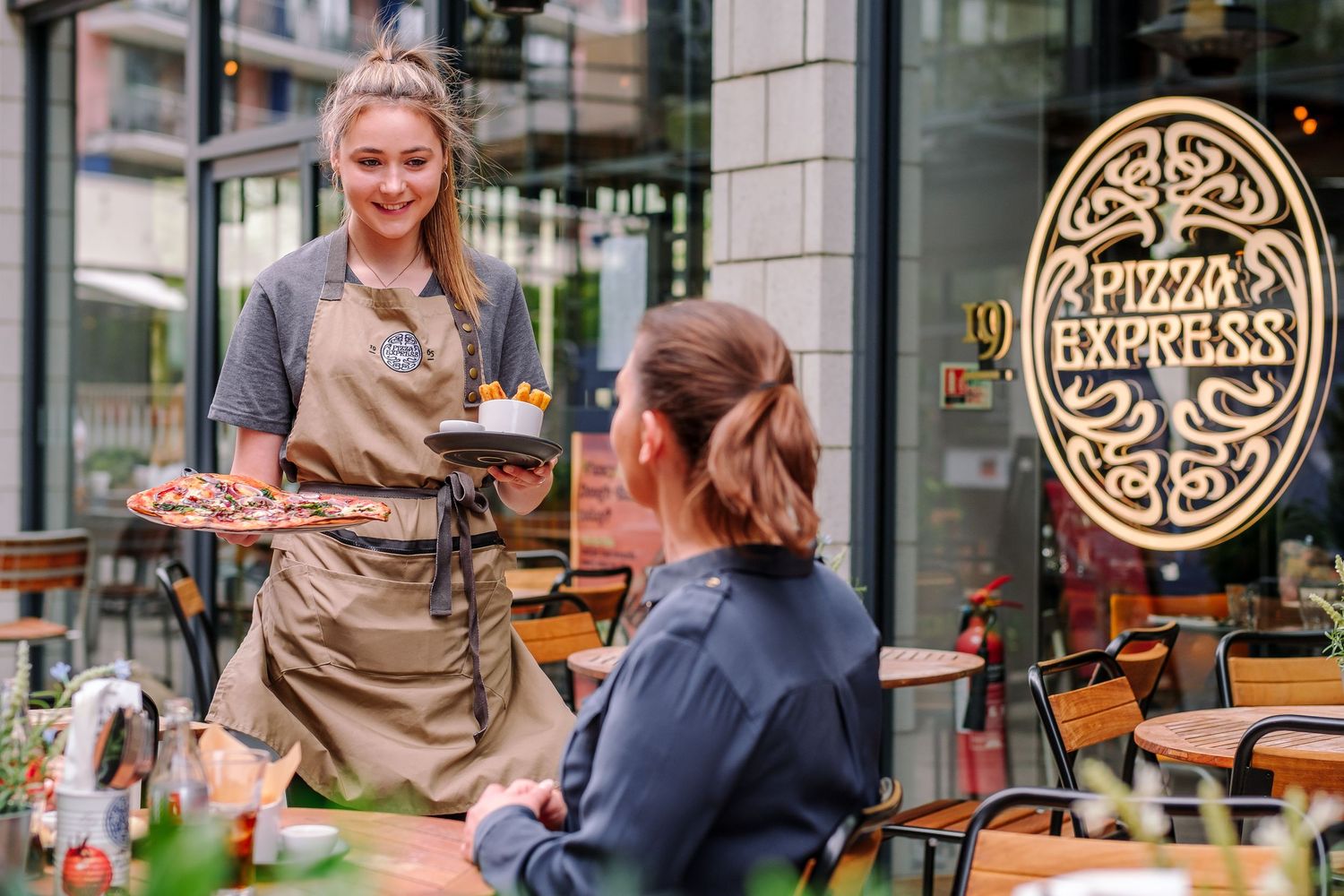 A server in a tan apron takes an order at an outdoor cafe with decorative signage.
