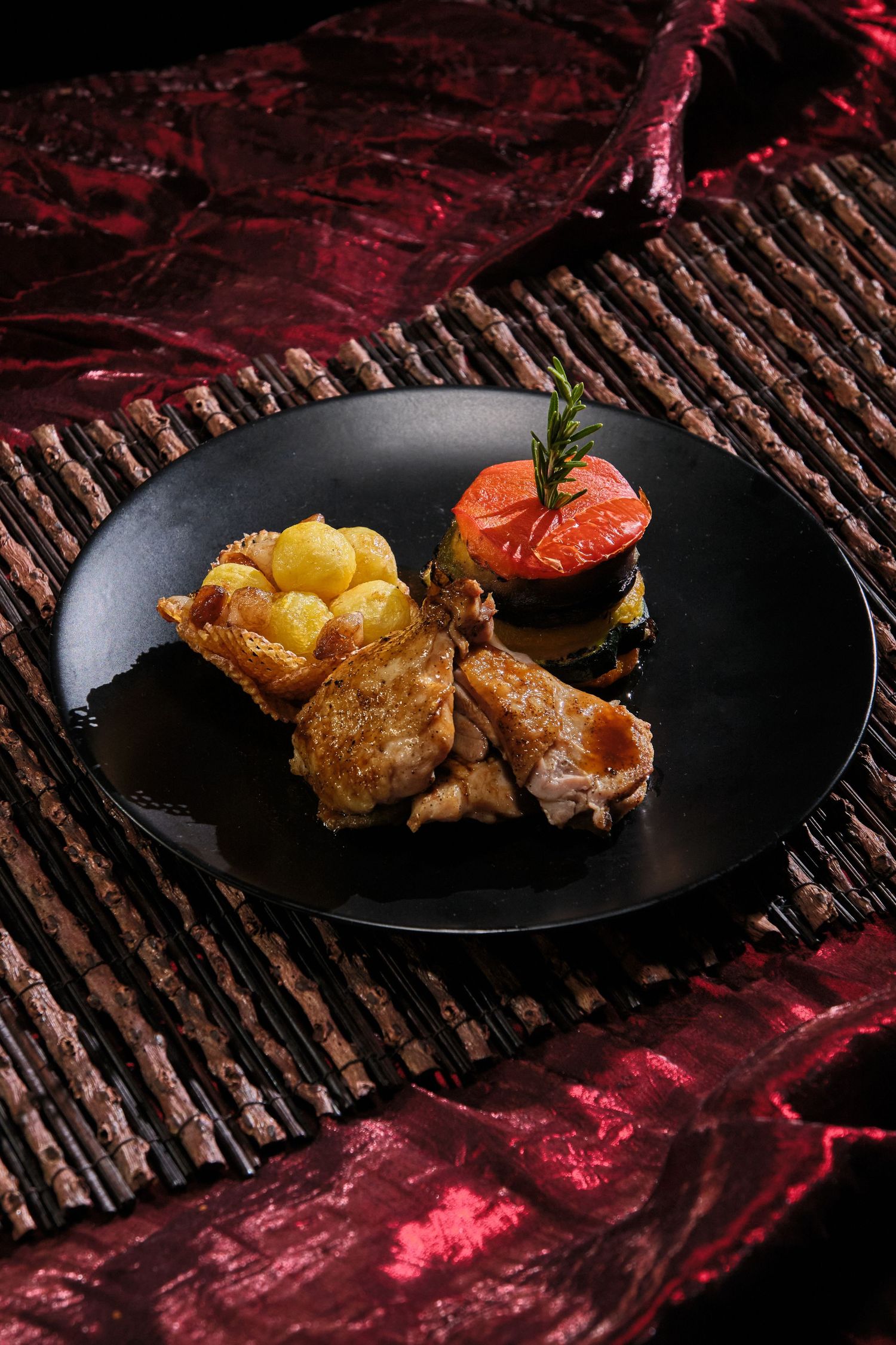 Elegant black plate with roasted meat, golden potatoes, and a stacked vegetable tower garnished with rosemary.