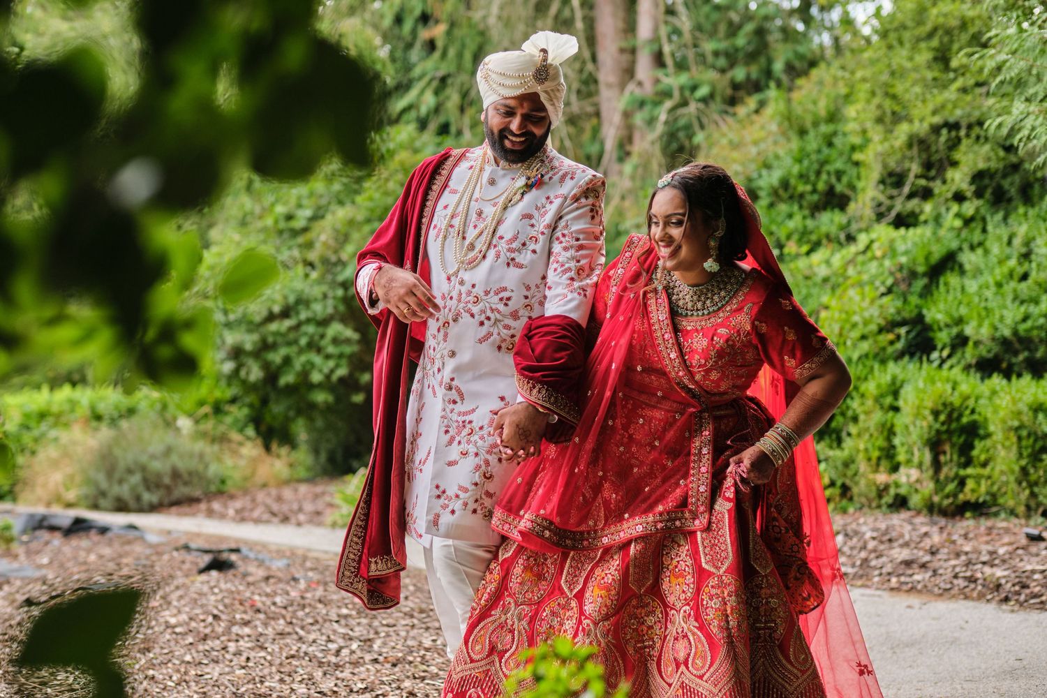 Couple in traditional South Asian wedding attire poses among lush green garden foliage.