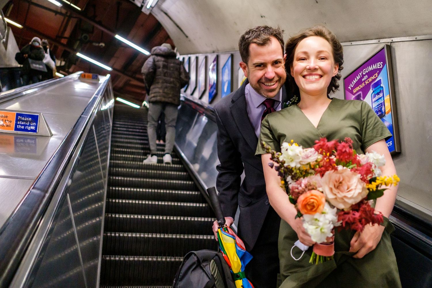 Happy couple running through a subway station holding colorful umbrellas and a bouquet of flowers.