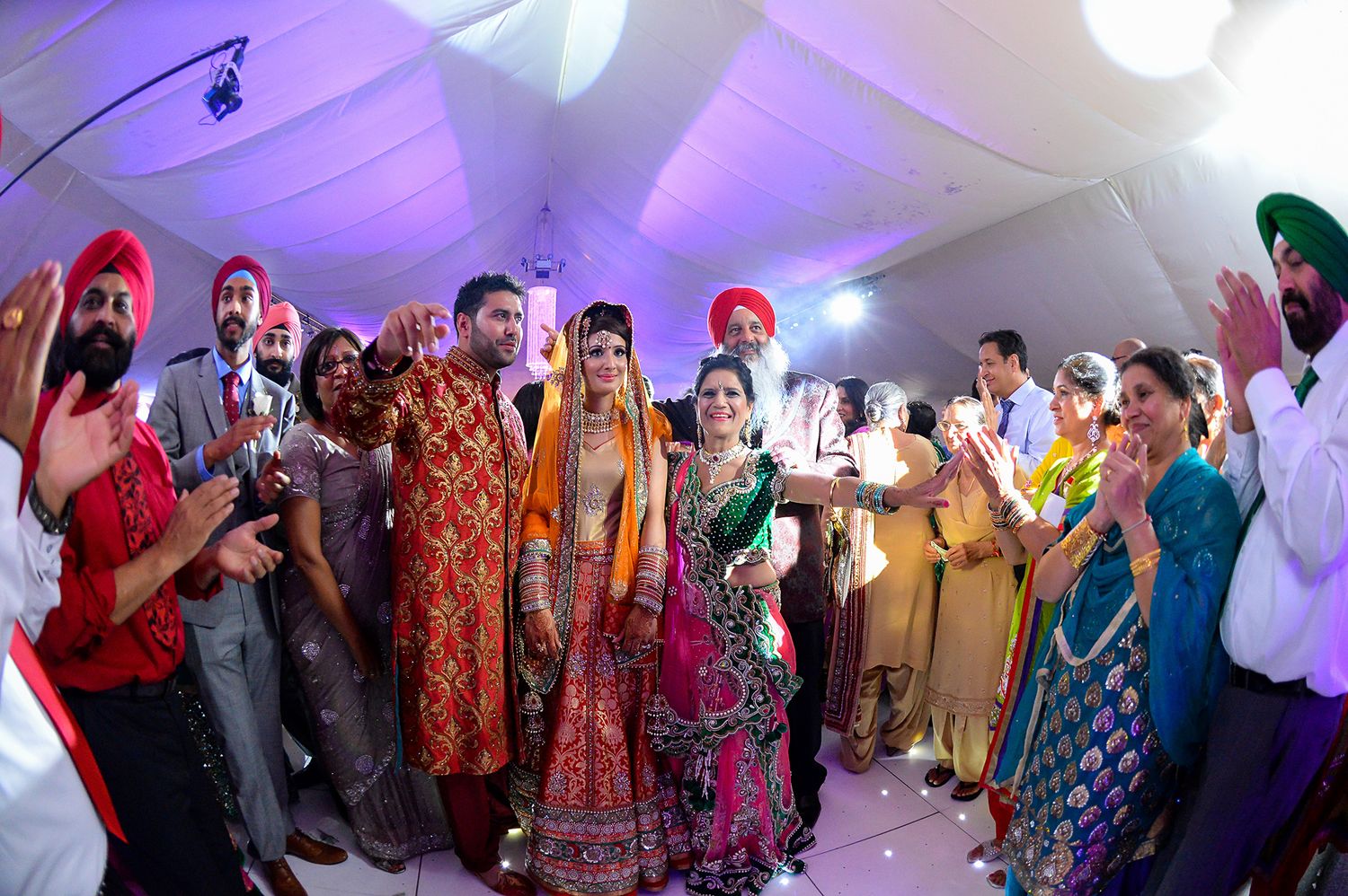 Colorful Sikh wedding celebration with guests dancing under purple-lit tent.
