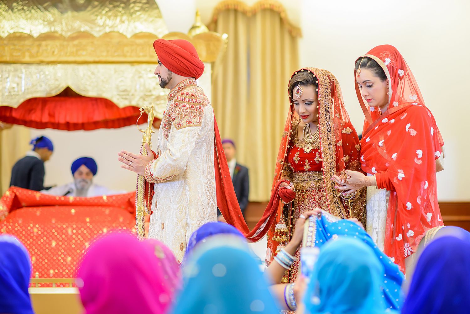 Guests in bright traditional dress gather for a Sikh wedding ceremony at an indoor venue.