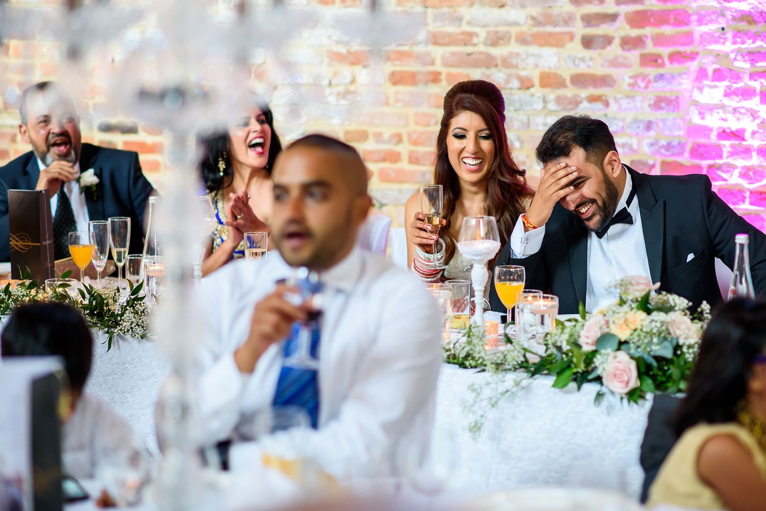 Wedding reception scene showing decorated tables and guests in a rustic brick-walled venue.