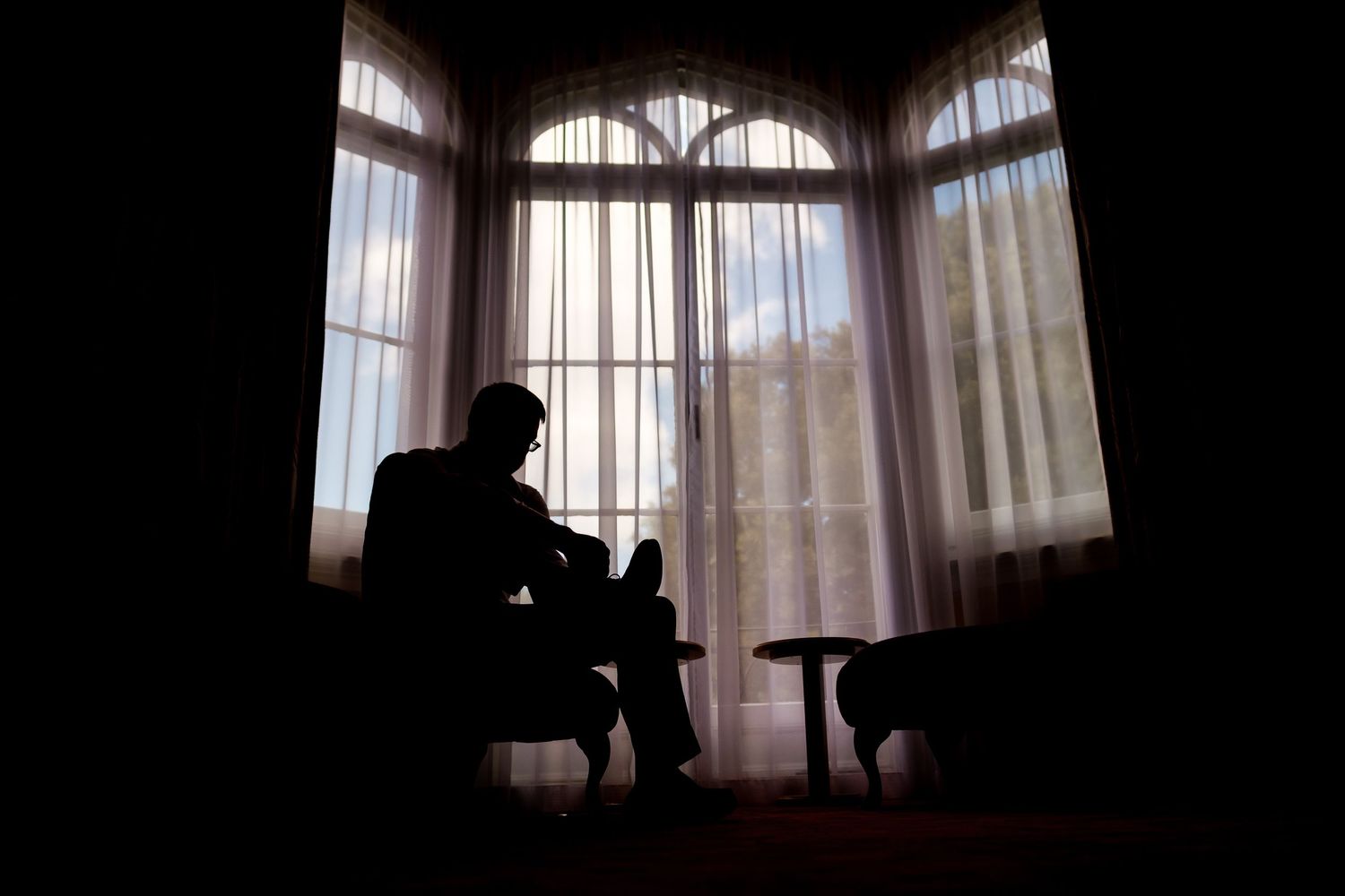 Silhouette of person sitting alone in chair by bay window with sheer curtains in dark moody room.