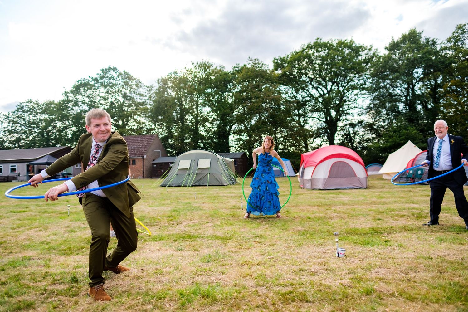 Wedding guests enjoy festivities on a grassy field with camping tents in background.