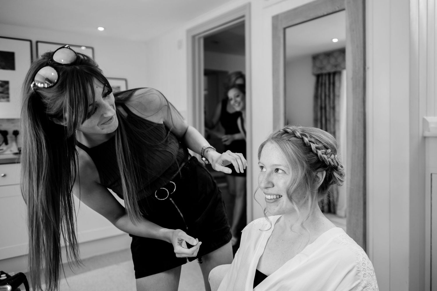 A stylist works on a bride's hair during wedding preparations in a bright white dressing room.
