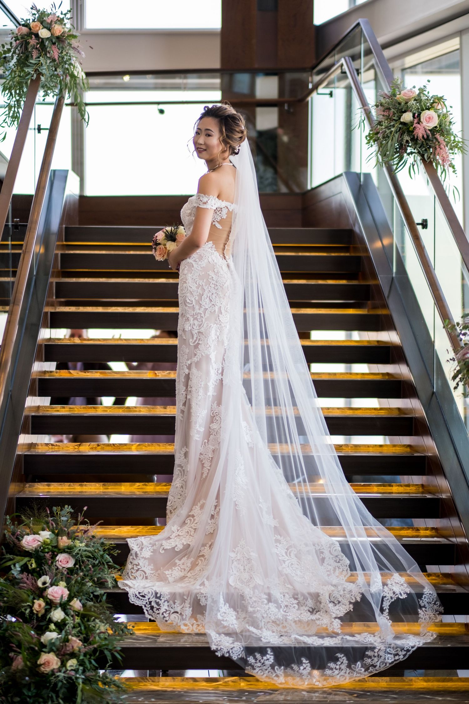 Elegant wedding dress with lace details and flowing veil displayed on wooden staircase decorated with floral arrangements.