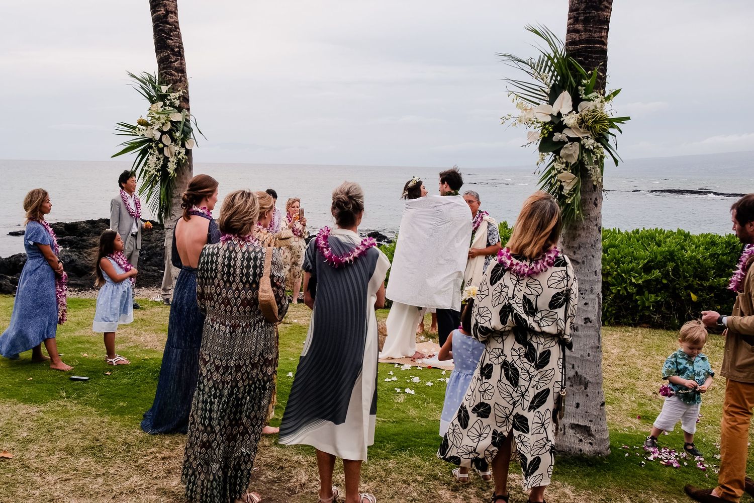 Wedding ceremony guests standing outdoors under palm trees at a beachfront location.