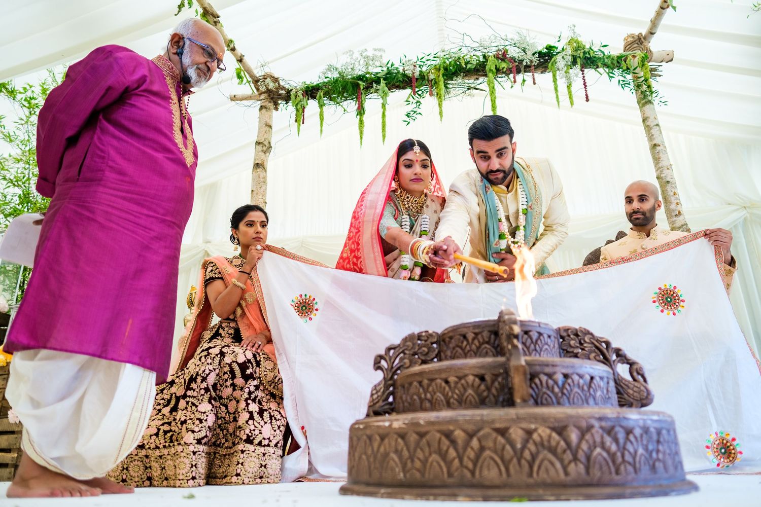 Traditional Indian wedding ceremony with decorative cake display under floral mandap with white fabric backdrop.