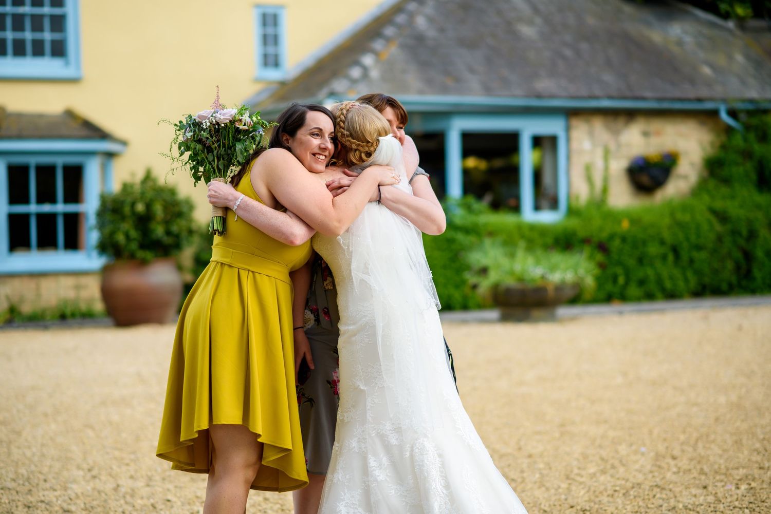 Bride in white dress embraces guests wearing yellow and navy outside yellow building.