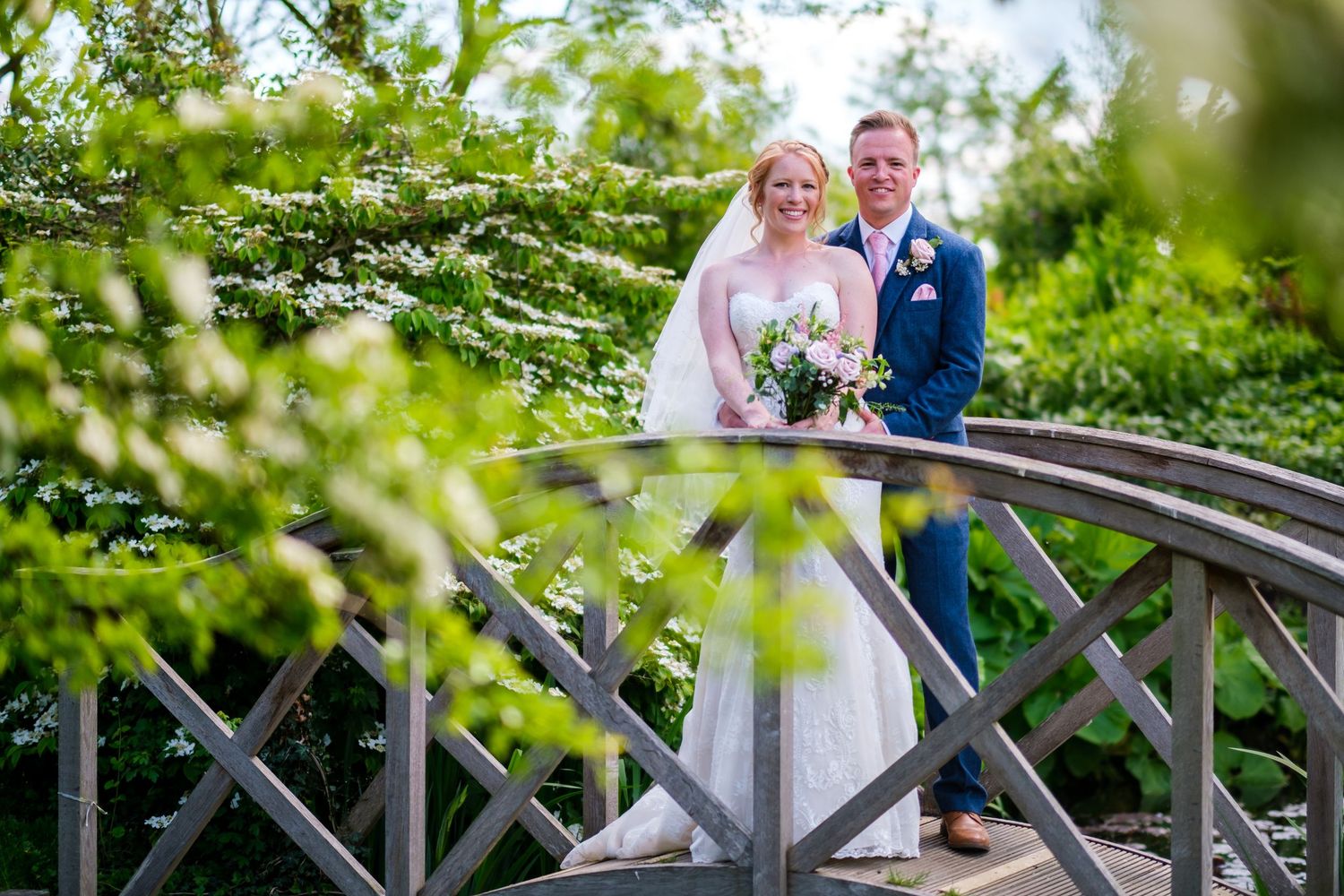 Wedding couple poses on a rustic wooden bridge surrounded by lush summer greenery.