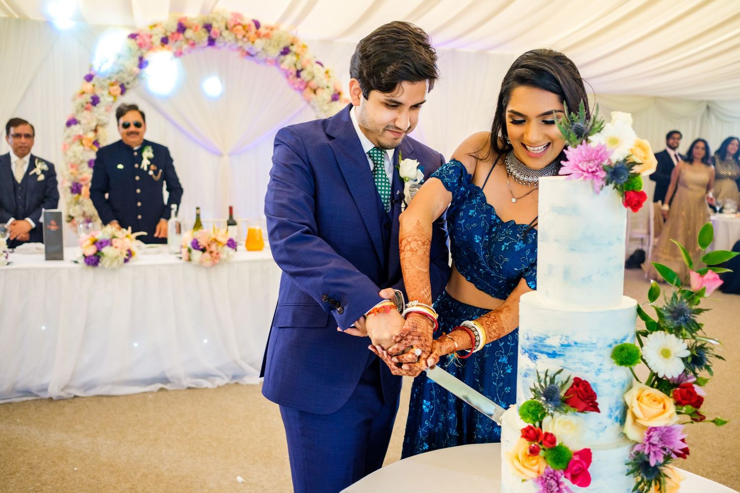 Couple in matching blue attire cut their wedding cake together under floral arch at reception.