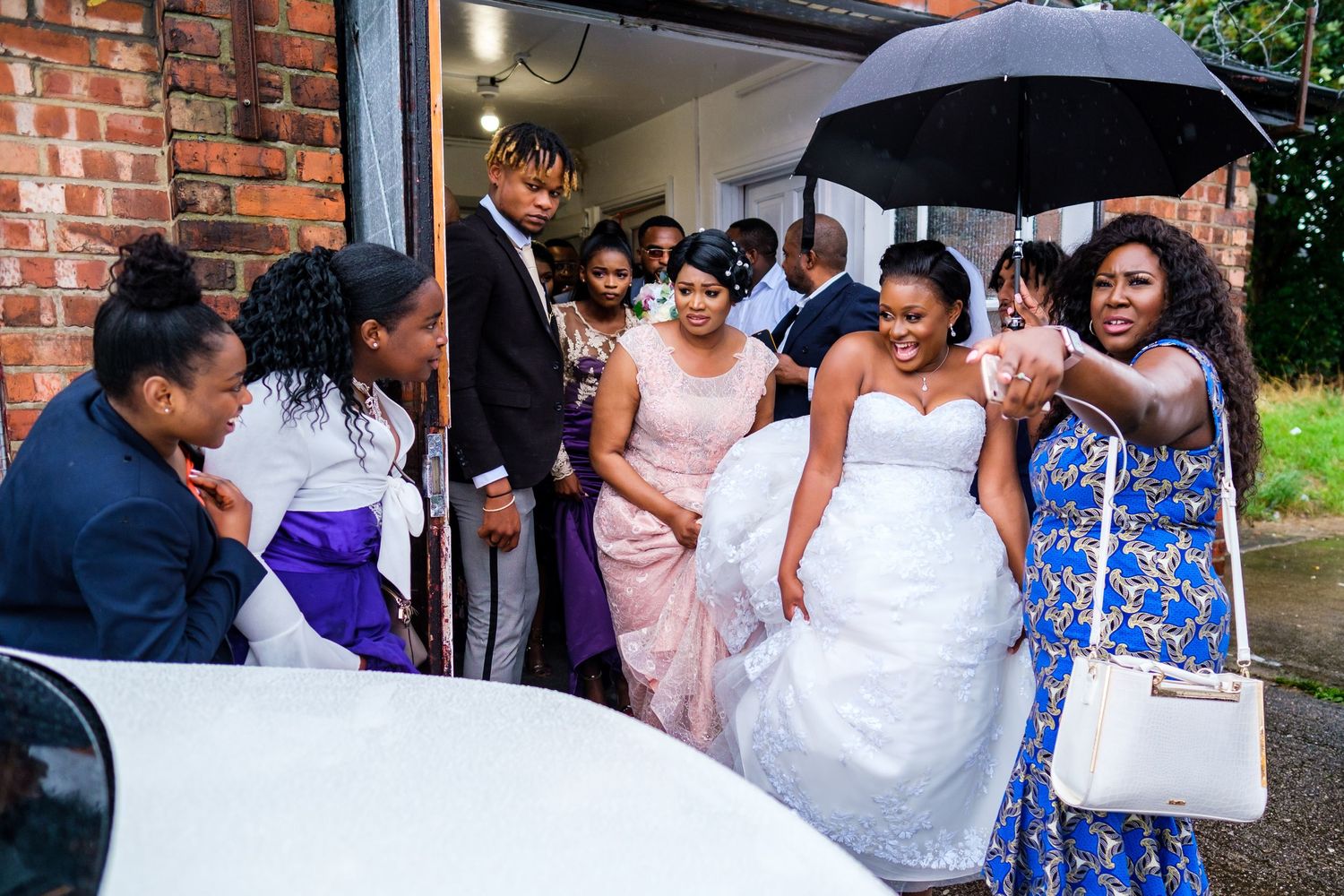 Wedding party gathered under umbrellas outside brick building in formal attire.