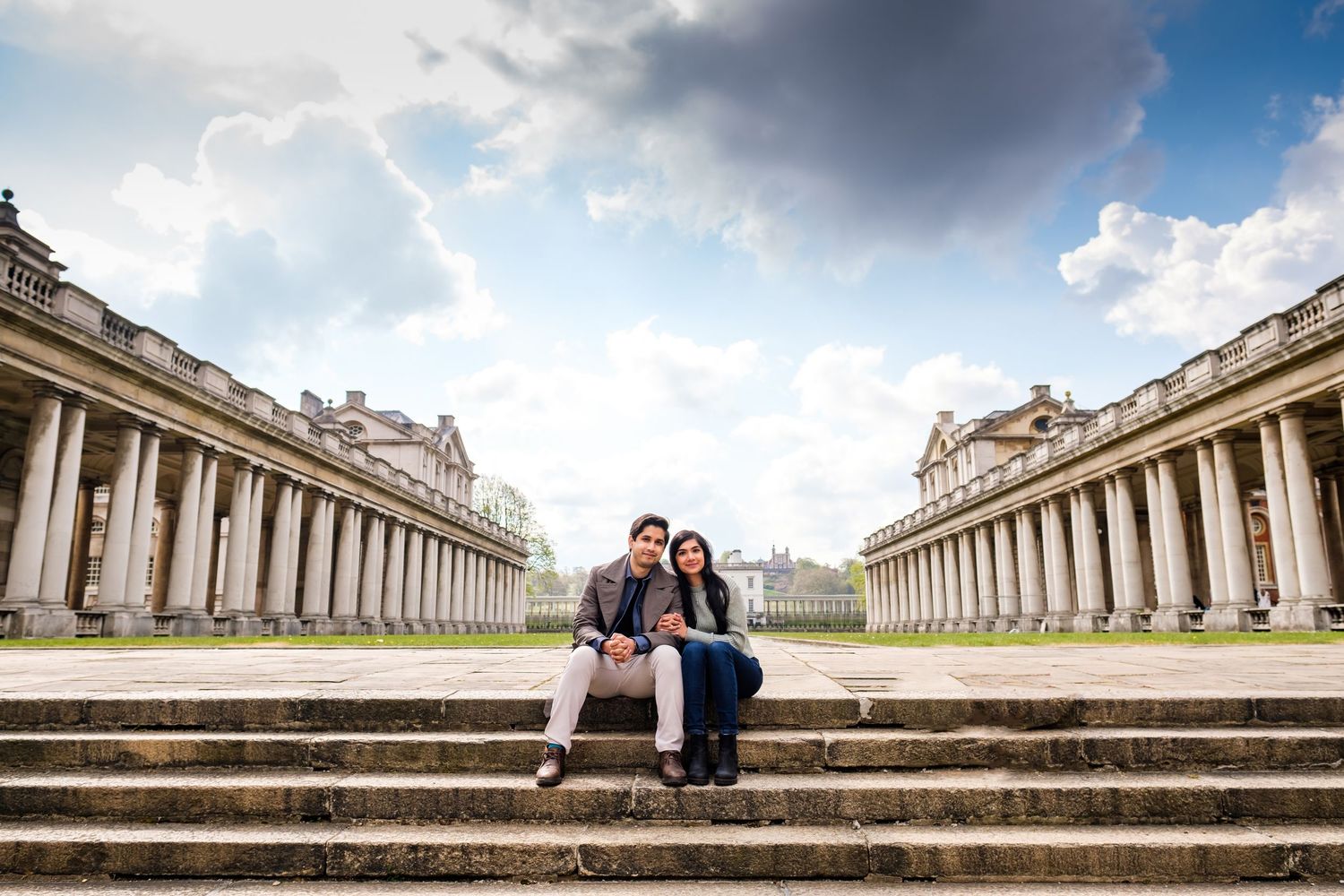 A couple sits on the grand steps of a historic European building with ornate classical architecture under dramatic clouds.