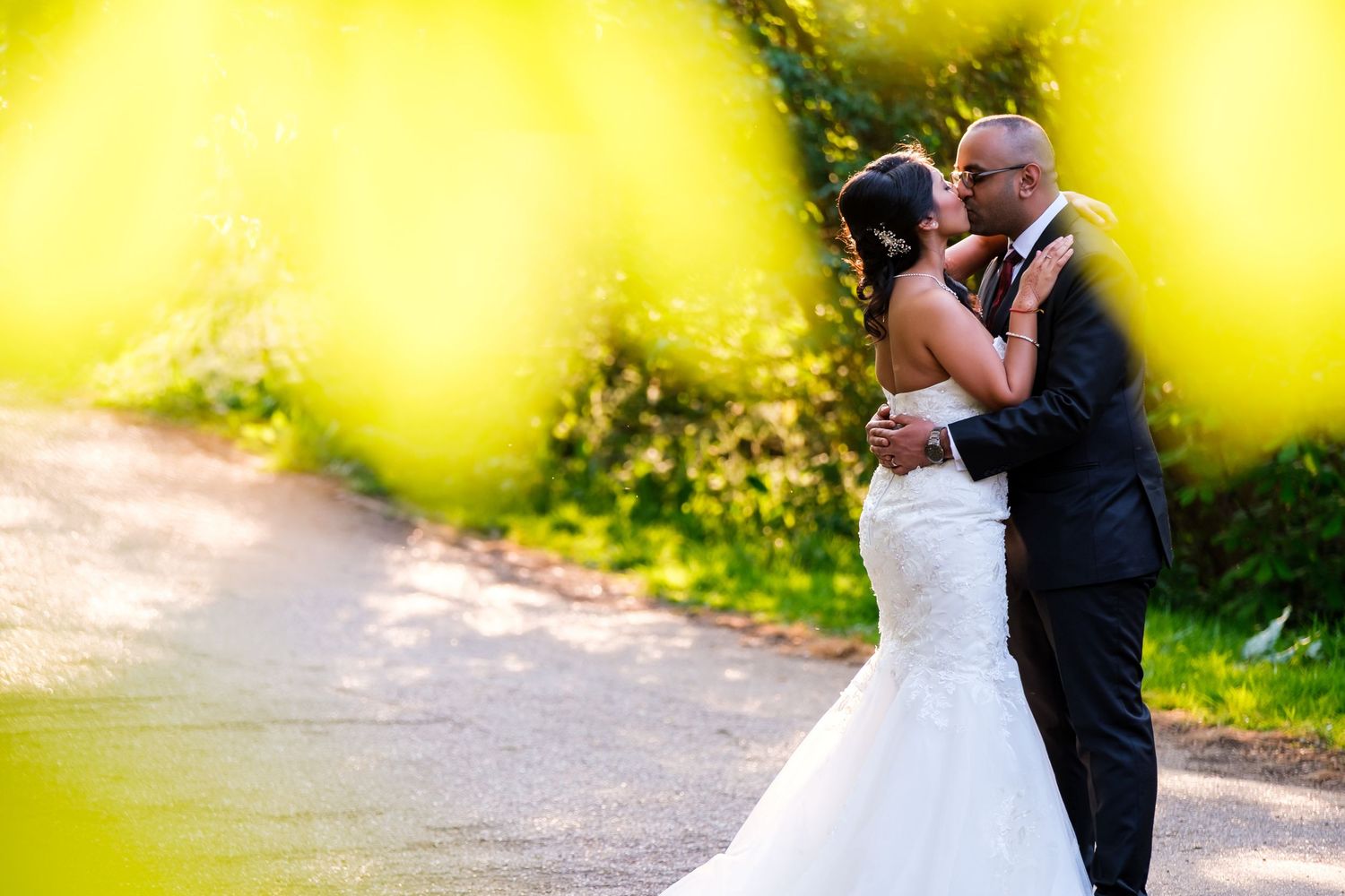 A romantic wedding moment captured through yellow flowers with a bride in white lace gown embracing on a garden path.