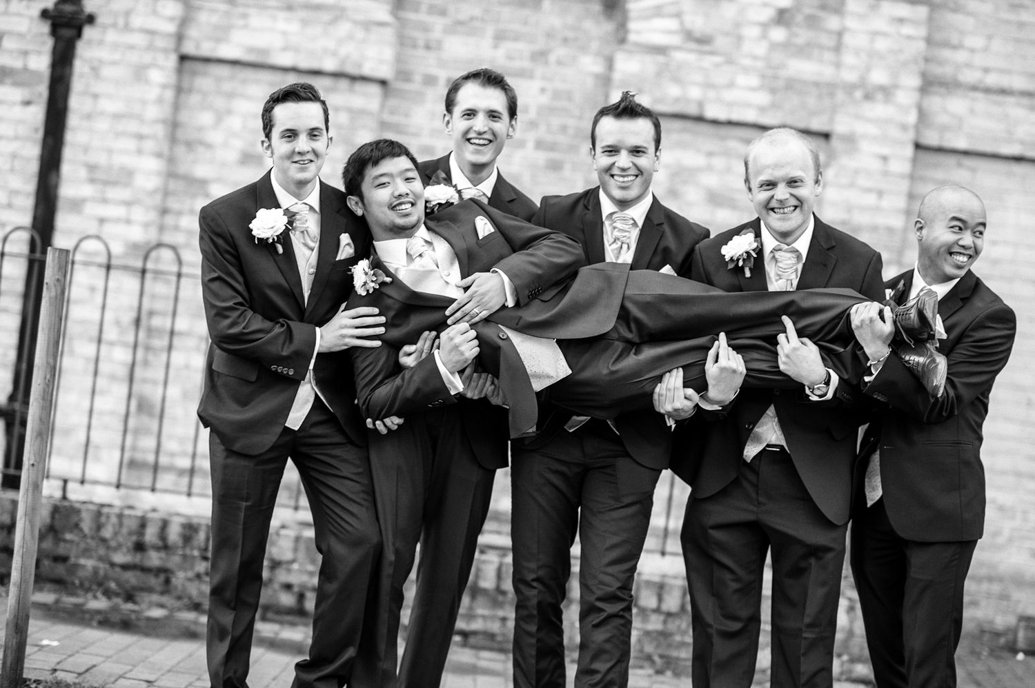 Groomsmen lift and carry groom in playful black and white portrait outside church.