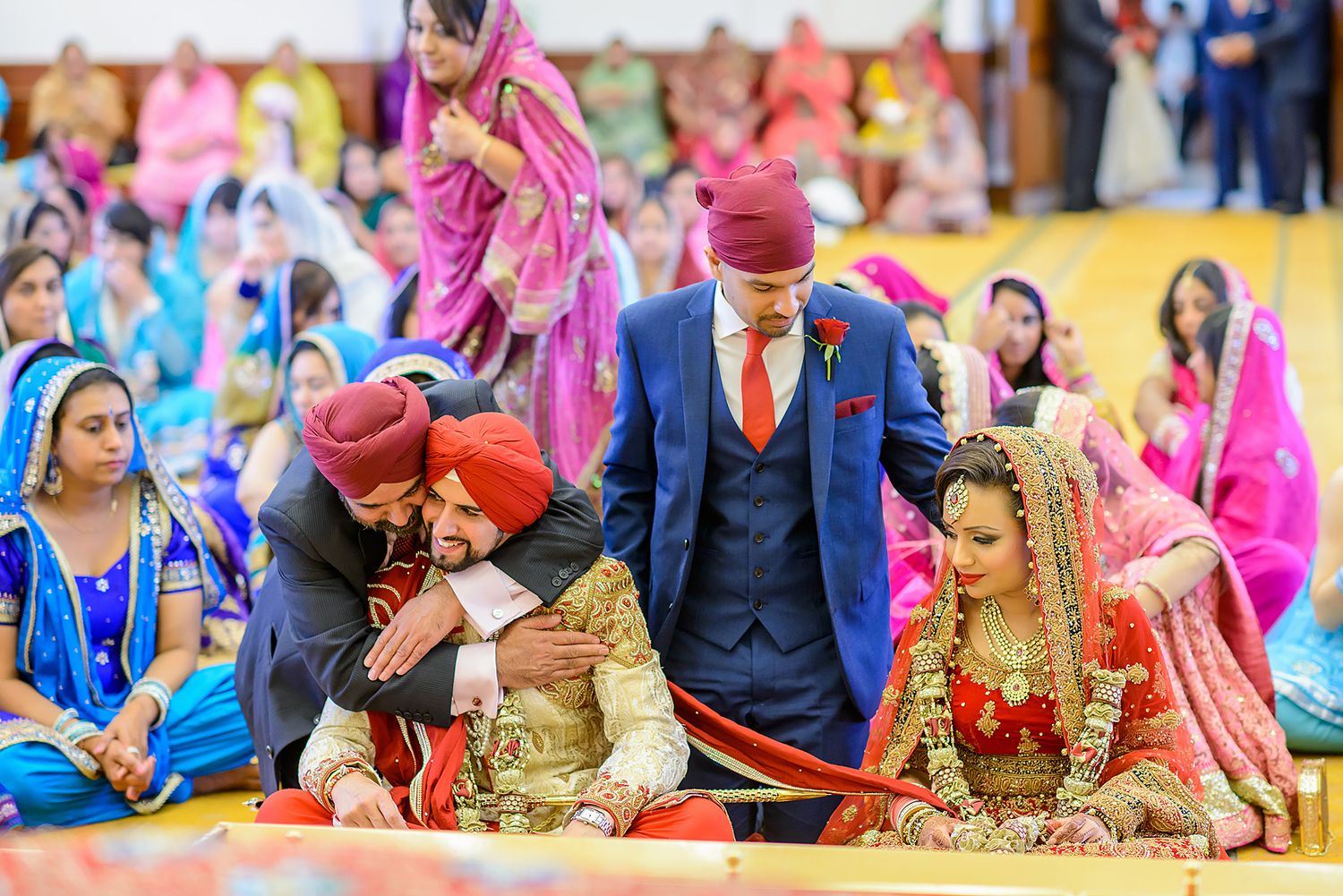Wedding ceremony participants seated on floor in traditional Sikh wedding attire with red and blue colors.