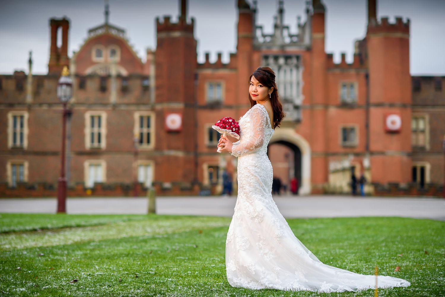 Bride in lace gown photographed against backdrop of grand historic building with red brick facade.