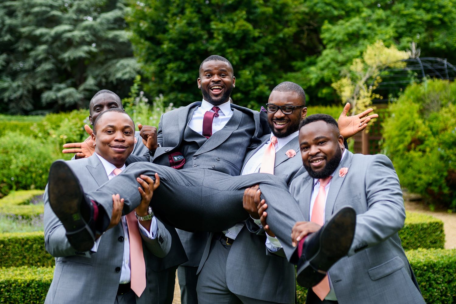 Groomsmen in gray suits and pink ties pose playfully for group photos outdoors.