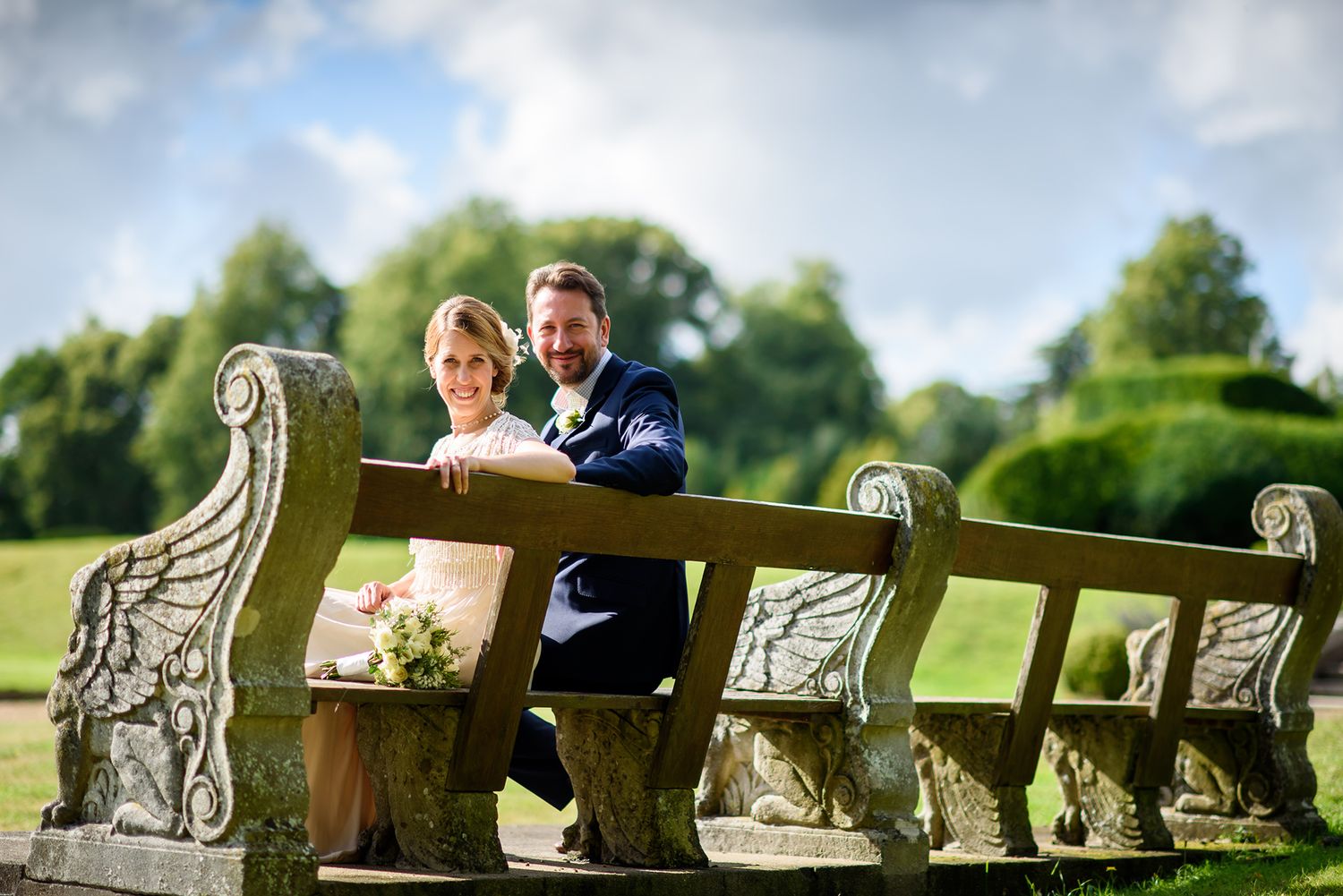 Wedding couple seated on ornate stone bench in garden setting.