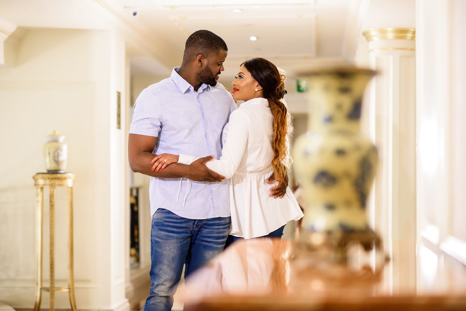 A romantic scene unfolds in a sunlit hallway with cream colored walls and classic architecture.