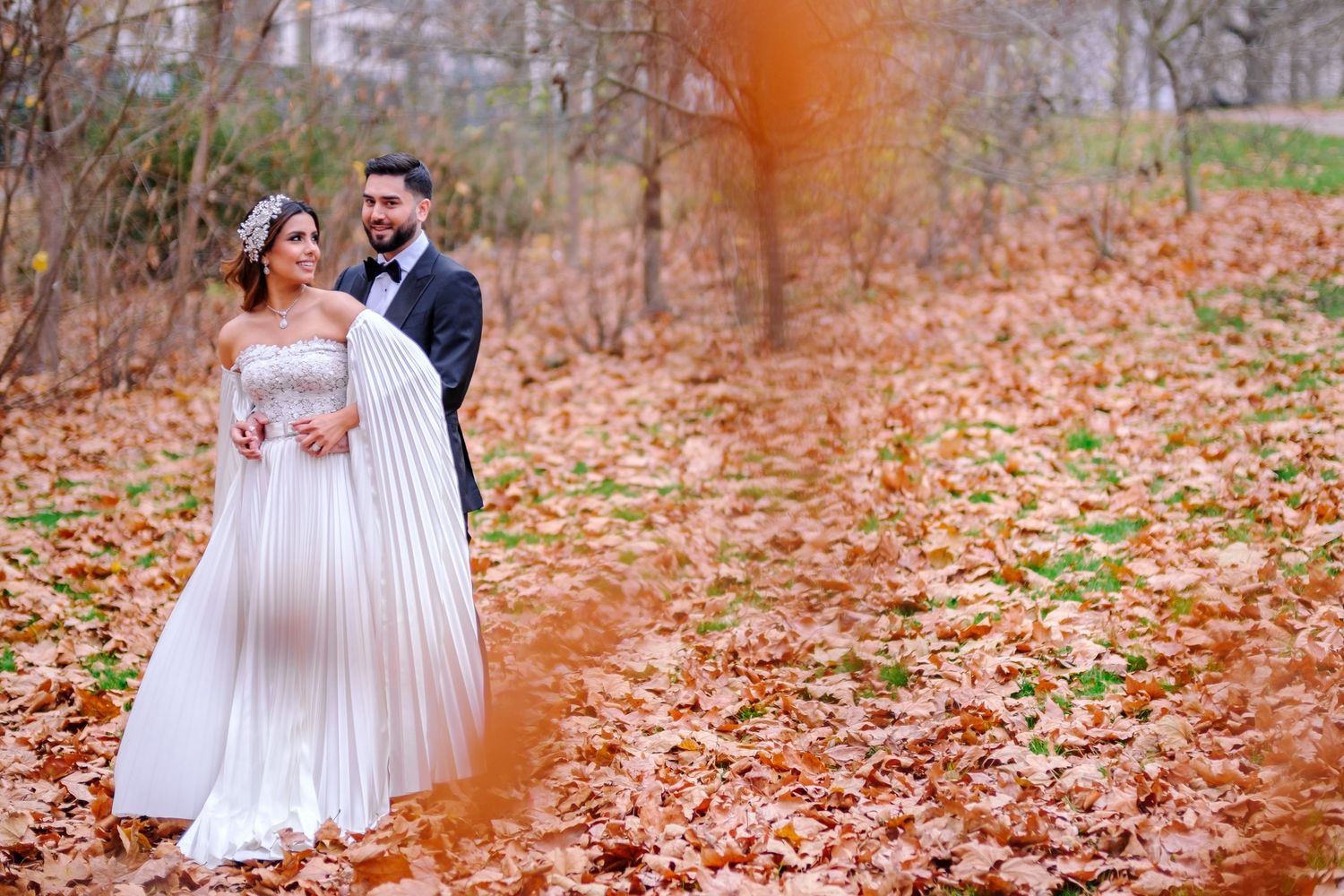 A couple in white wedding attire walks through a scenic autumn forest filled with fallen orange leaves.