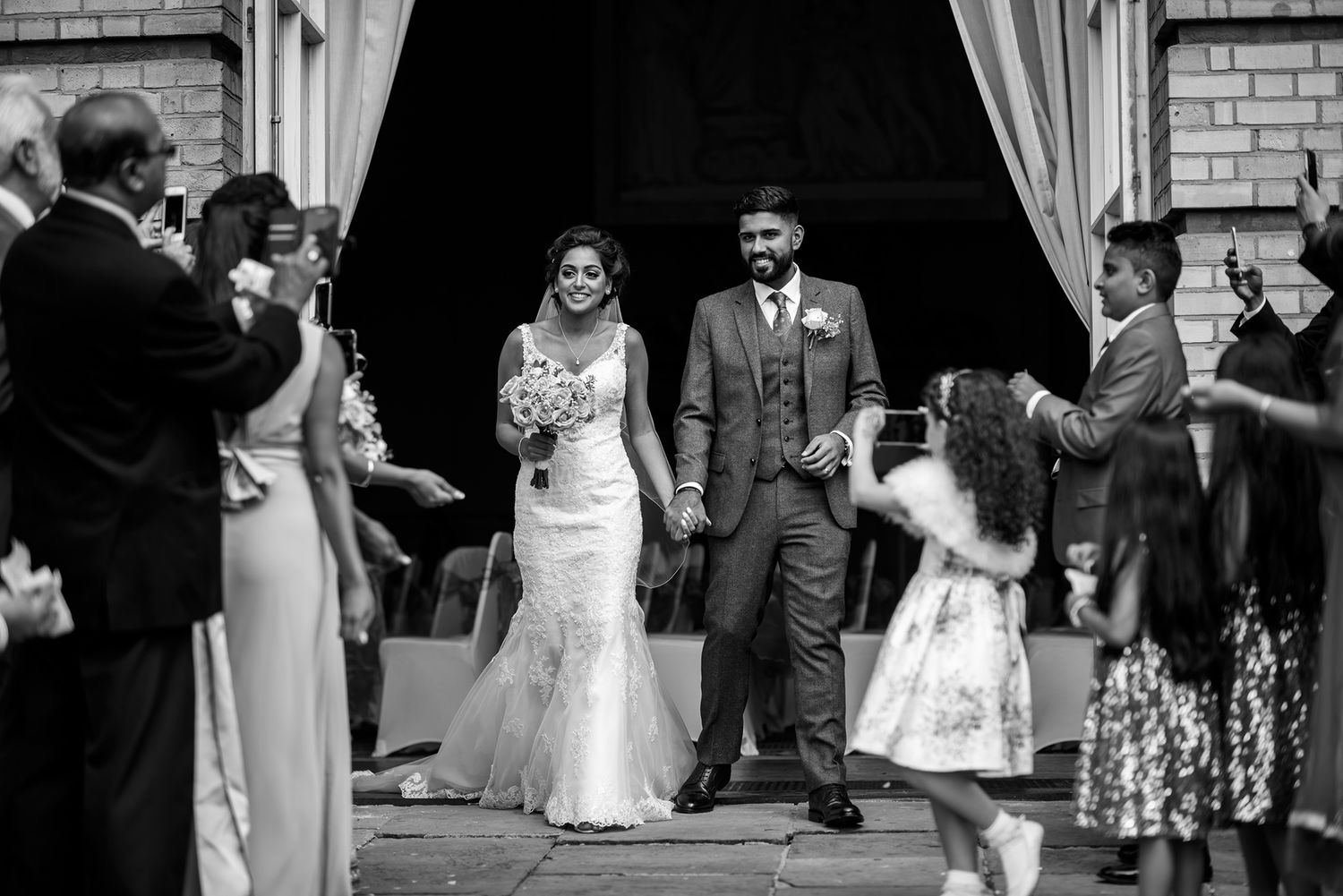 Wedding group poses together in front of brick building with white curtains framing the entrance.
