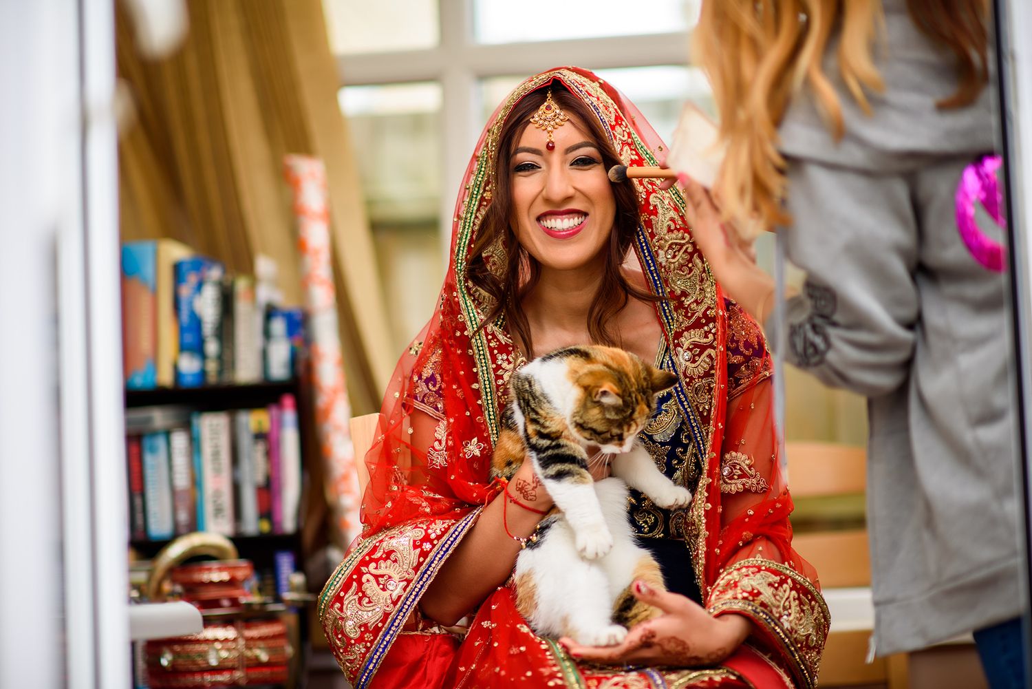 A bride in traditional red wedding attire holds a small white and black cat during a celebration.