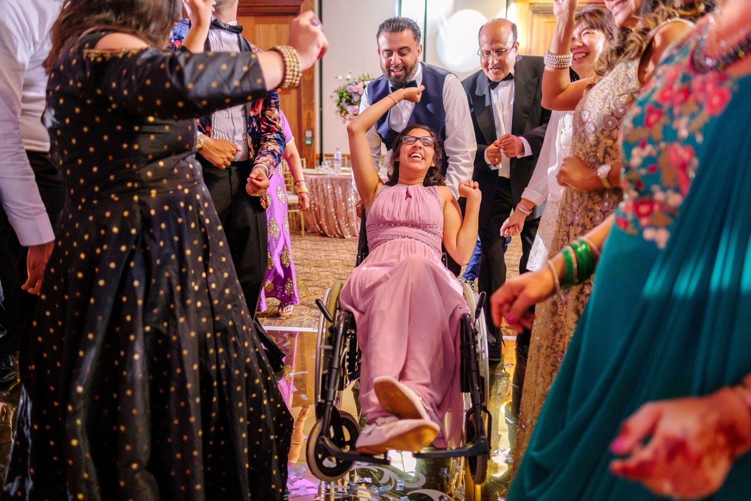 Wedding celebration scene with guests dancing around someone in a pink dress seated in a wheelchair.
