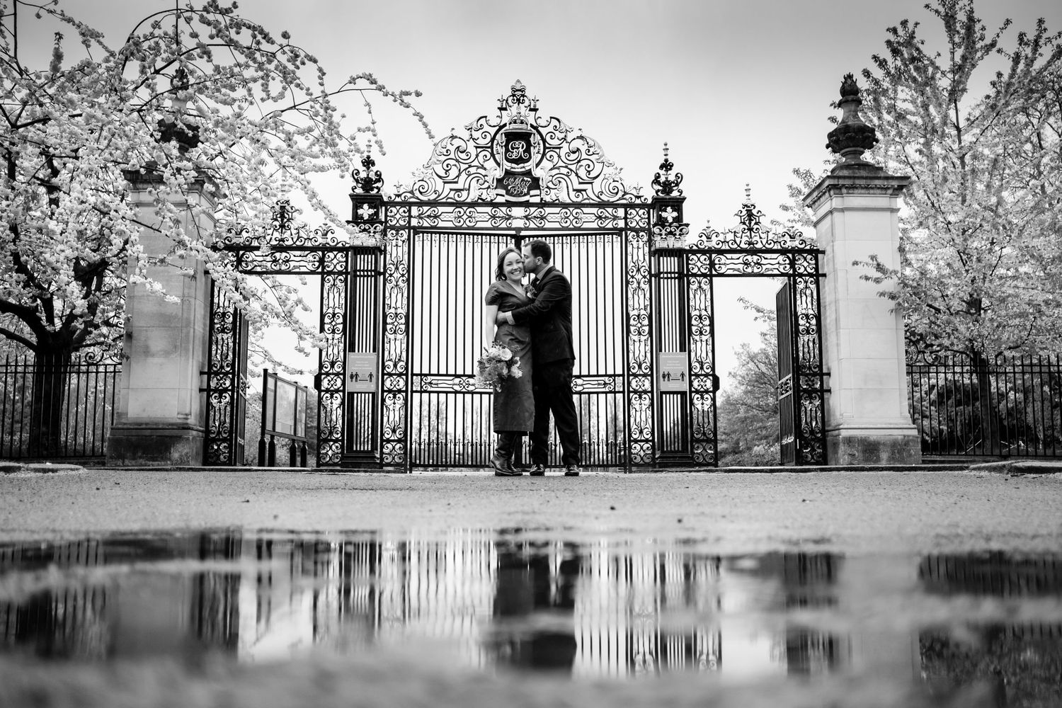 A black and white panoramic view of an ornate iron gate entrance with people silhouetted against the background.
