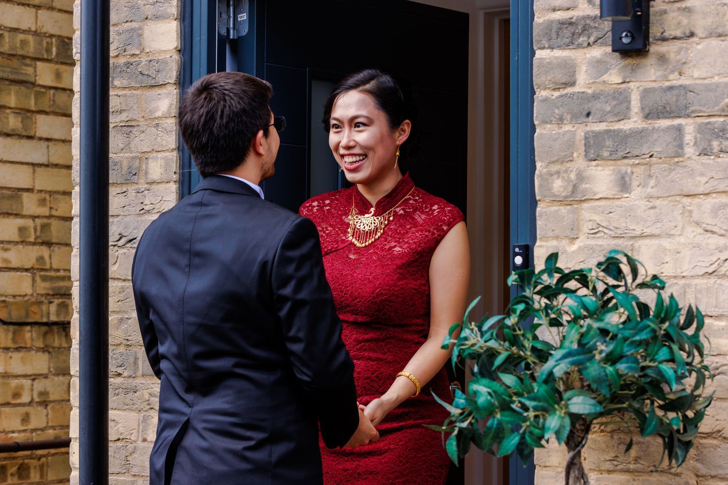 Couple in red qipao dress and dark suit standing in front of brick building entrance.