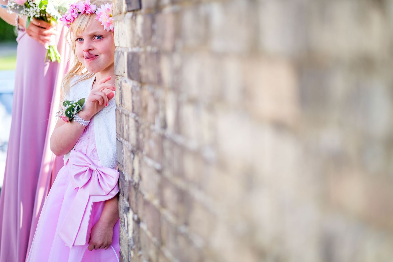 Child in pink dress with floral crown peeks around stone wall corner at outdoor event.
