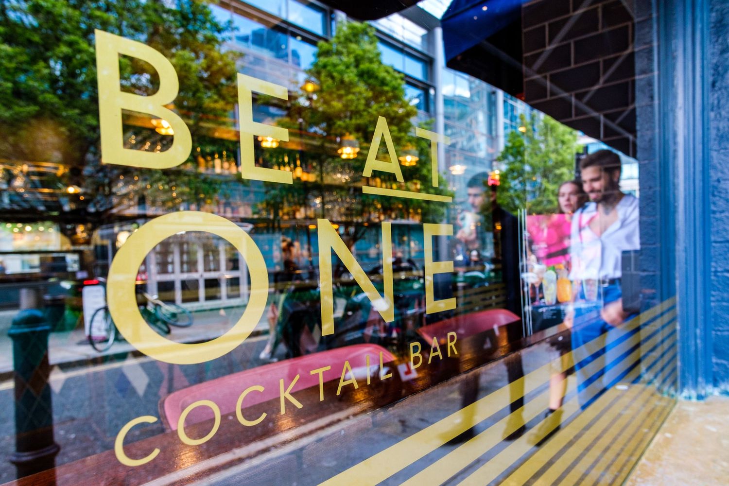 Storefront window with gold lettering spelling 'BE' and 'ONE' with cocktail bar signage.