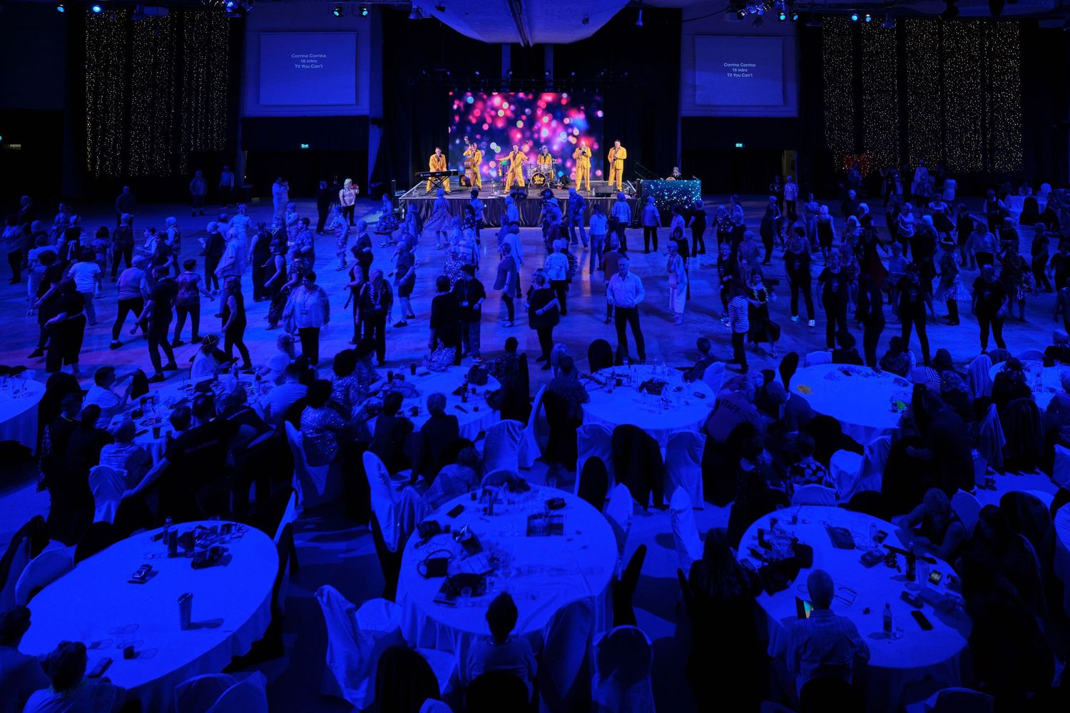 Large crowd of attendees seated at round tables in a blue-lit convention hall or banquet venue.