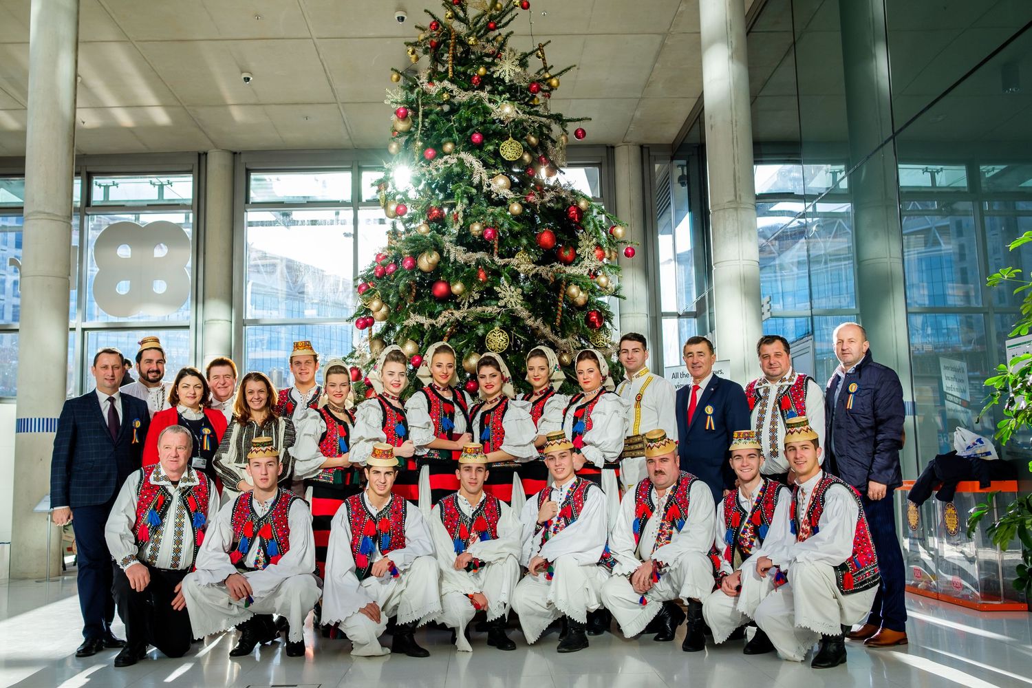 A group in traditional folk costumes poses together by a decorated Christmas tree in a modern building lobby.