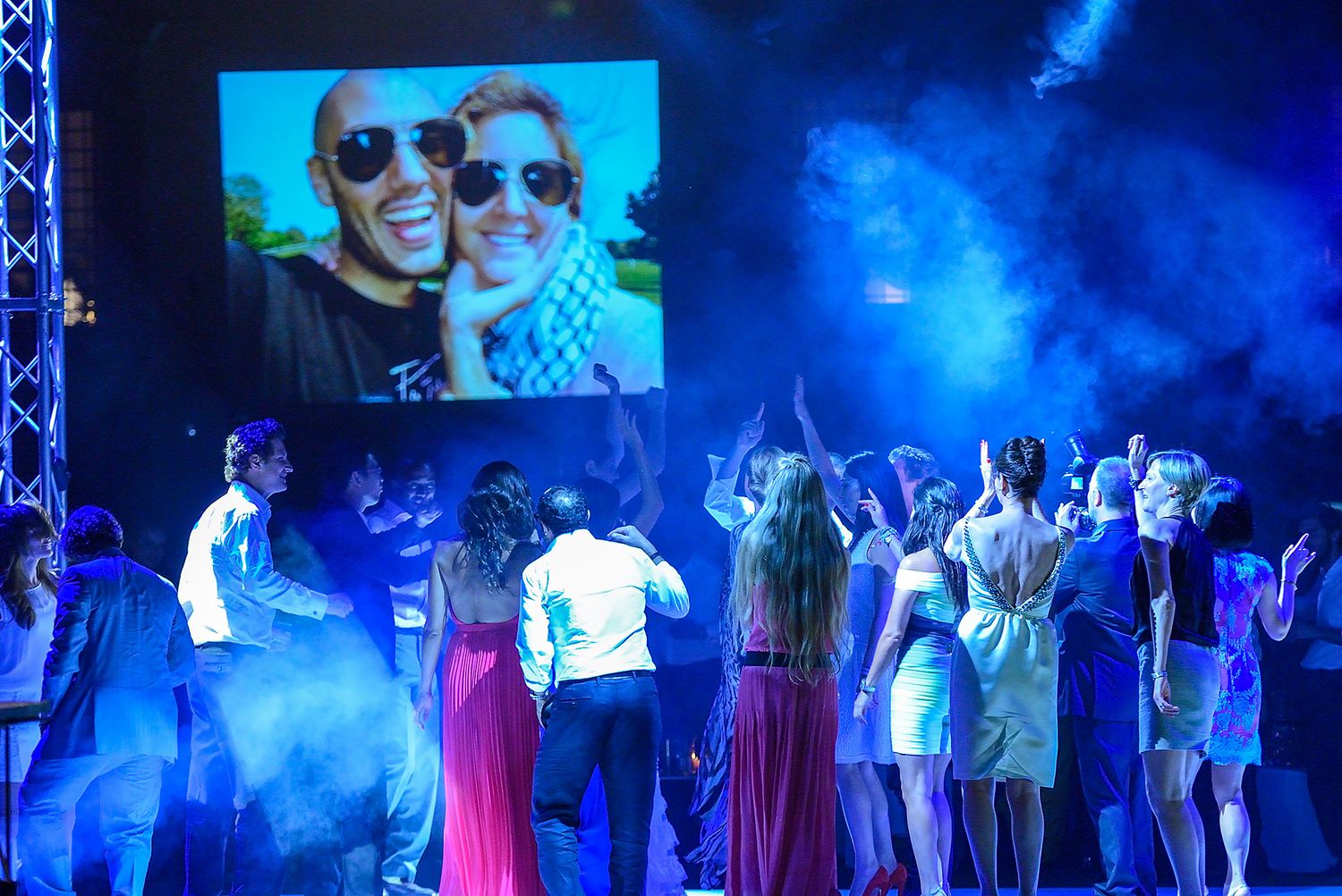 Crowd dances under blue stage lighting at a party with a large display screen showing selfies above.