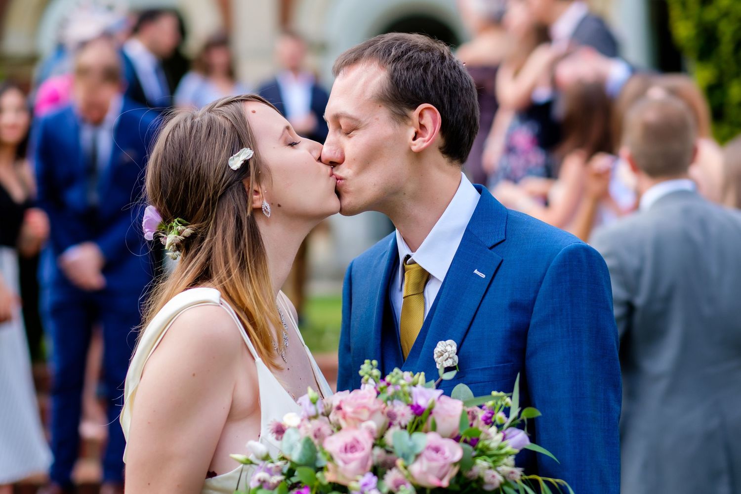 Newlyweds share a romantic kiss while guests celebrate in the background.