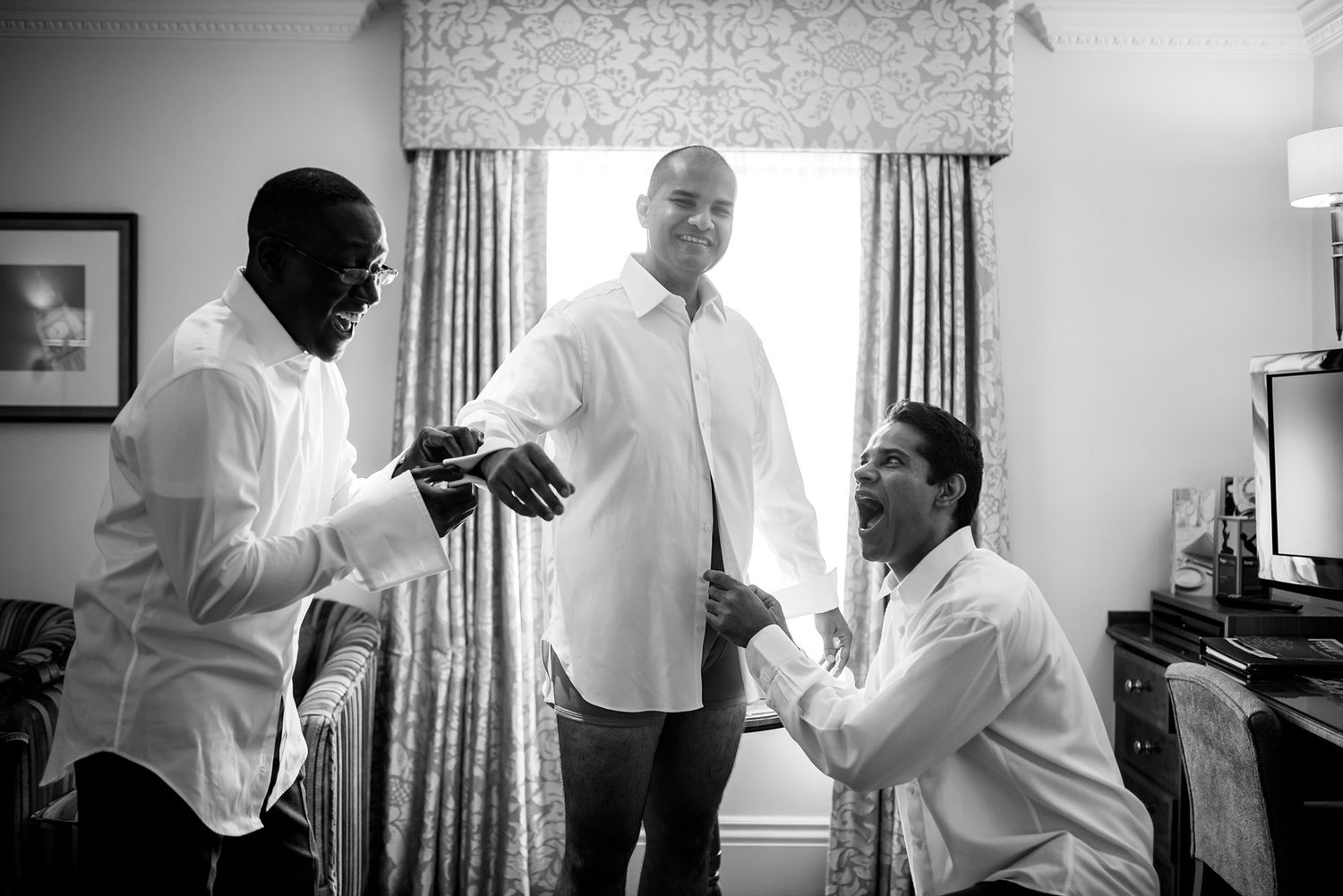 Wedding party members help the groom prepare in a sunlit room with white curtains.
