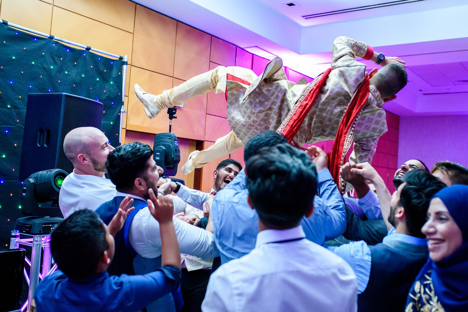 Wedding party guests dancing and celebrating with raised arms under purple ambient lighting.