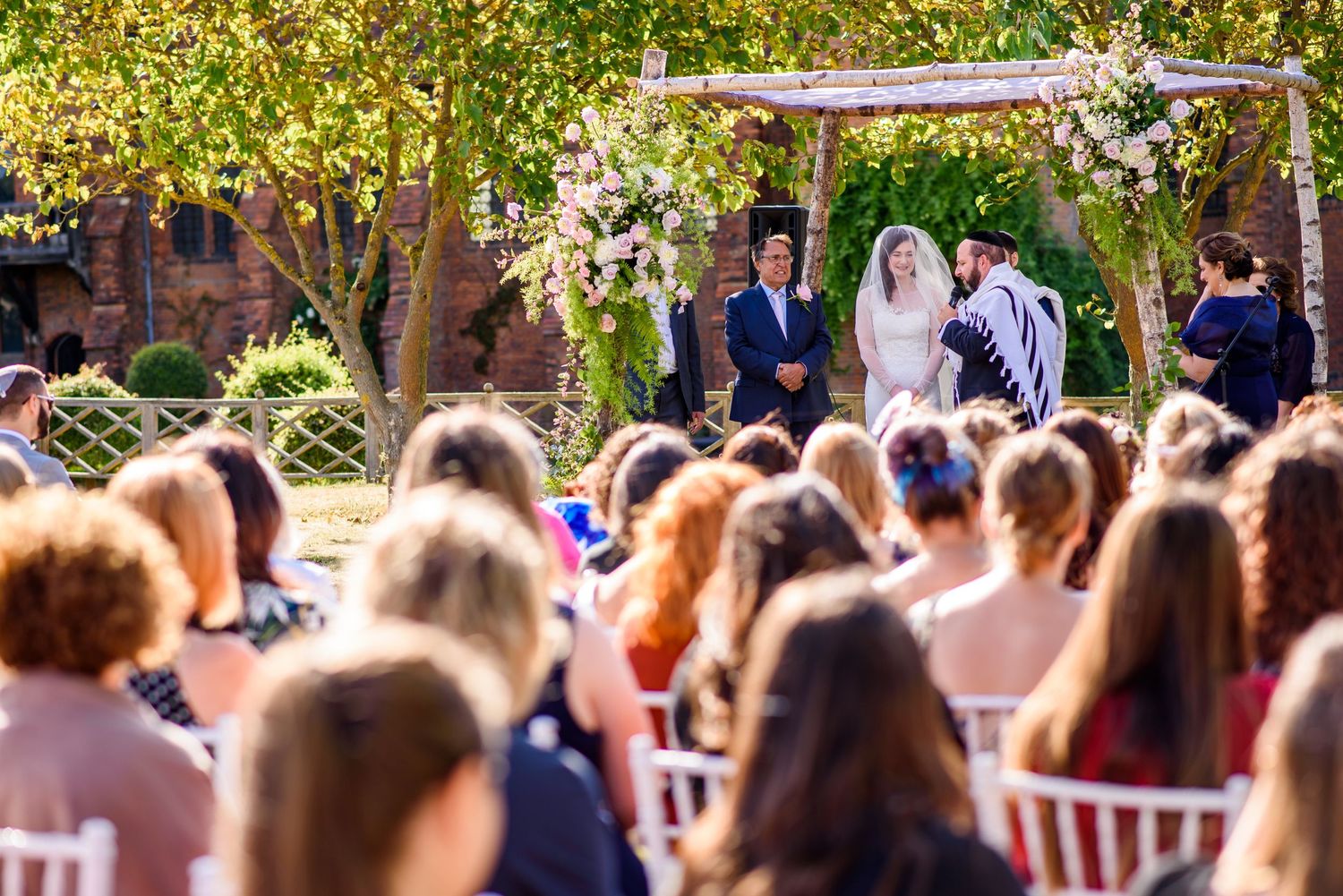 Wedding ceremony audience seated in white chairs outdoors with trees in the background.
