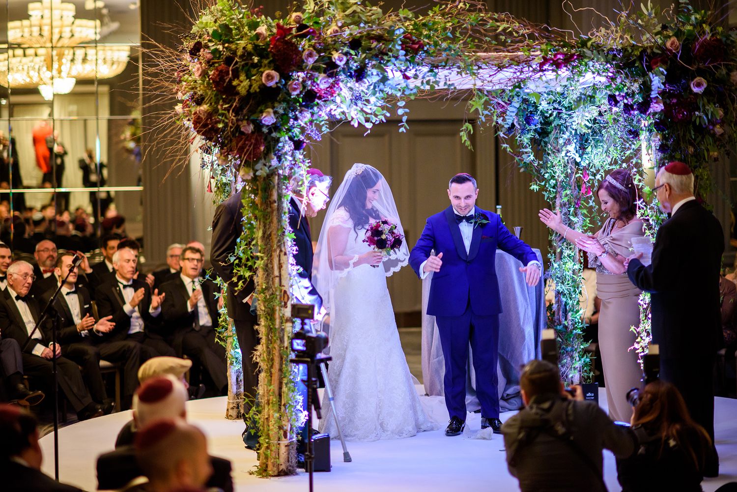Wedding ceremony moment under a stunning blue-lit floral arch with formal guests in attendance.